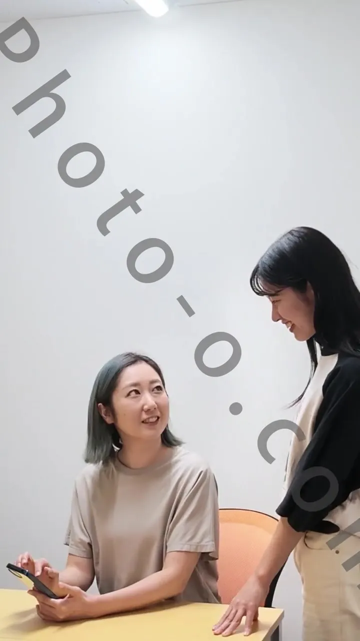 A seated performer holding a smartphone speaks with a standing performer in the production preview; Photo-o.com watermark retained